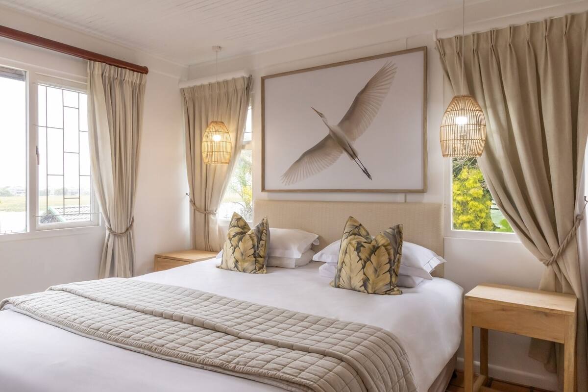 Un dormitorio con una cama grande, un cabecero de madera, una obra de arte enmarcada de un pájaro y vistas a la vegetación exterior.