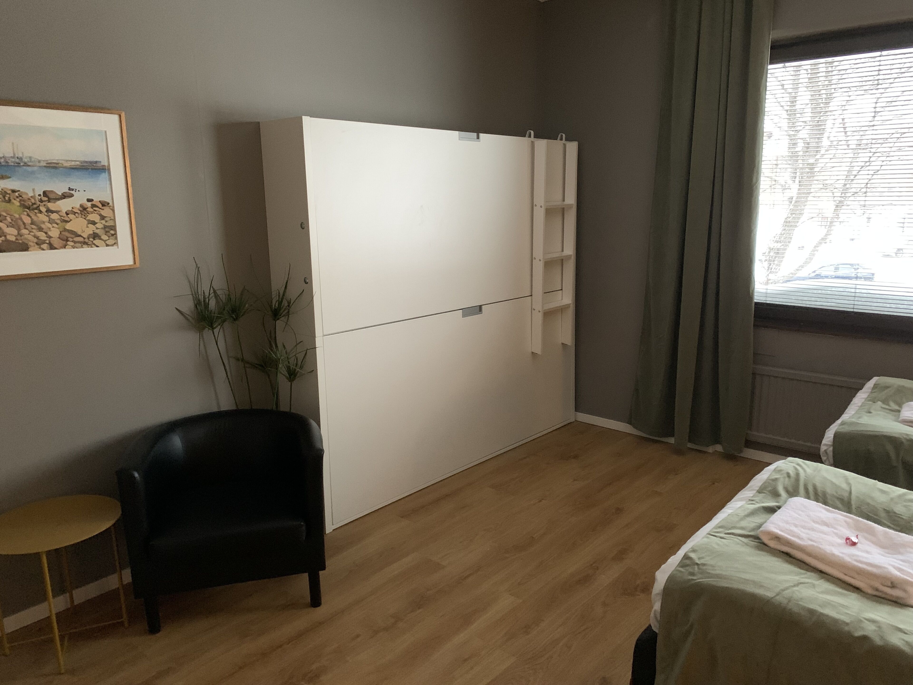 Ein Zimmer mit einem großen weißen Kleiderschrank, einem schwarzen Sessel, einem kleinen Tisch und zwei Betten mit grüner Bettwäsche.