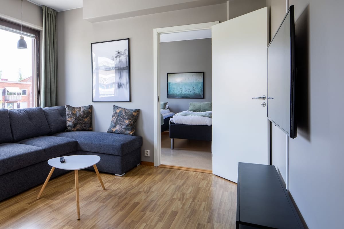 A modern living room with a grey sofa, a small round table, and a bed in the background.
