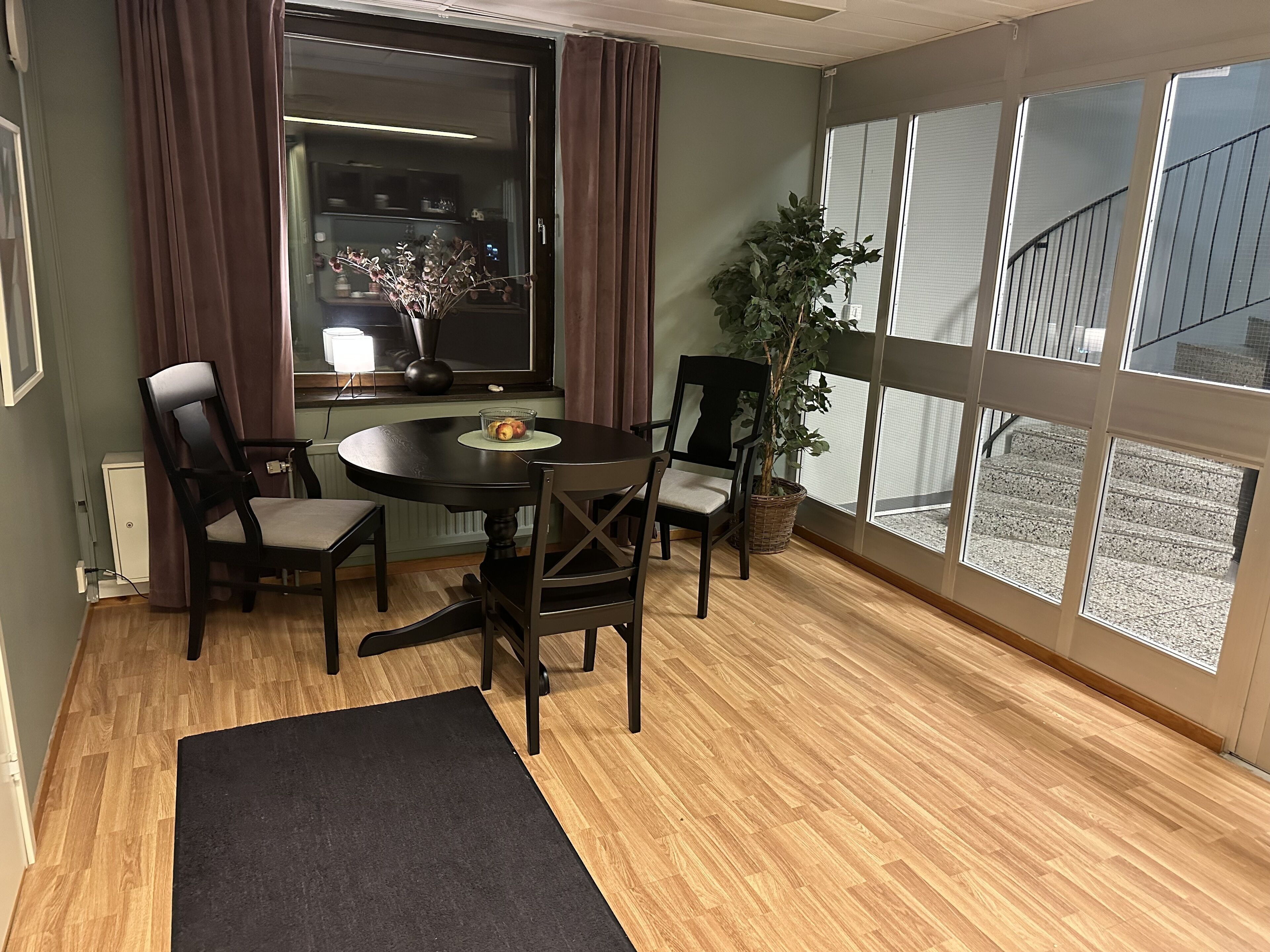 Een eetruimte met een ronde tafel, vier stoelen, een potplant en uitzicht op een gebouw.
