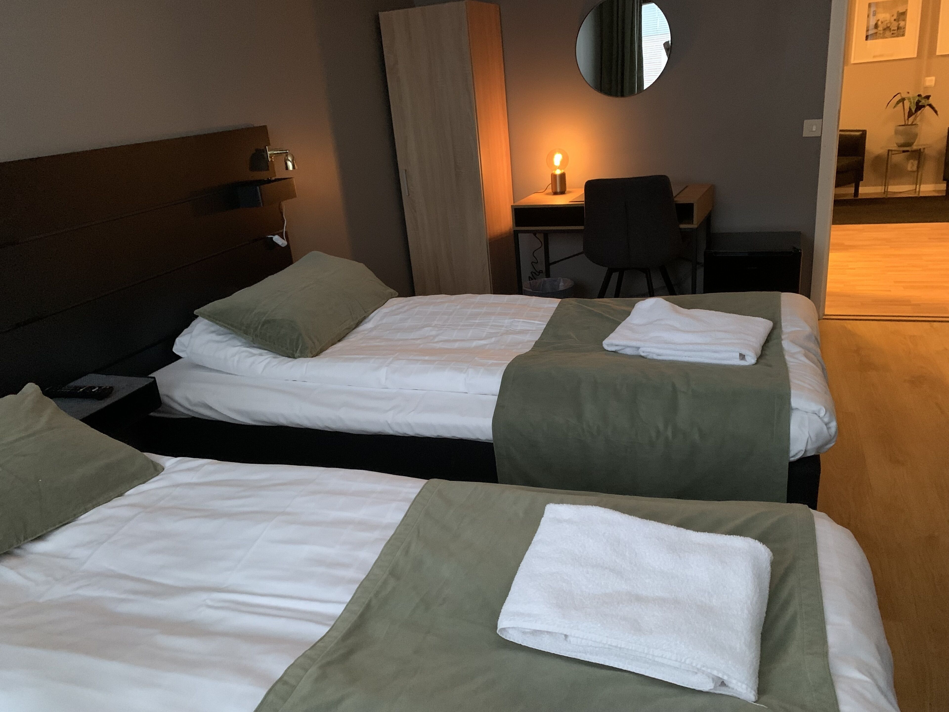 Habitación de hotel con dos camas, cada una con cubrecama verde y ropa de cama blanca. Hay un escritorio con una lámpara, una silla y un espejo redondo en la pared.