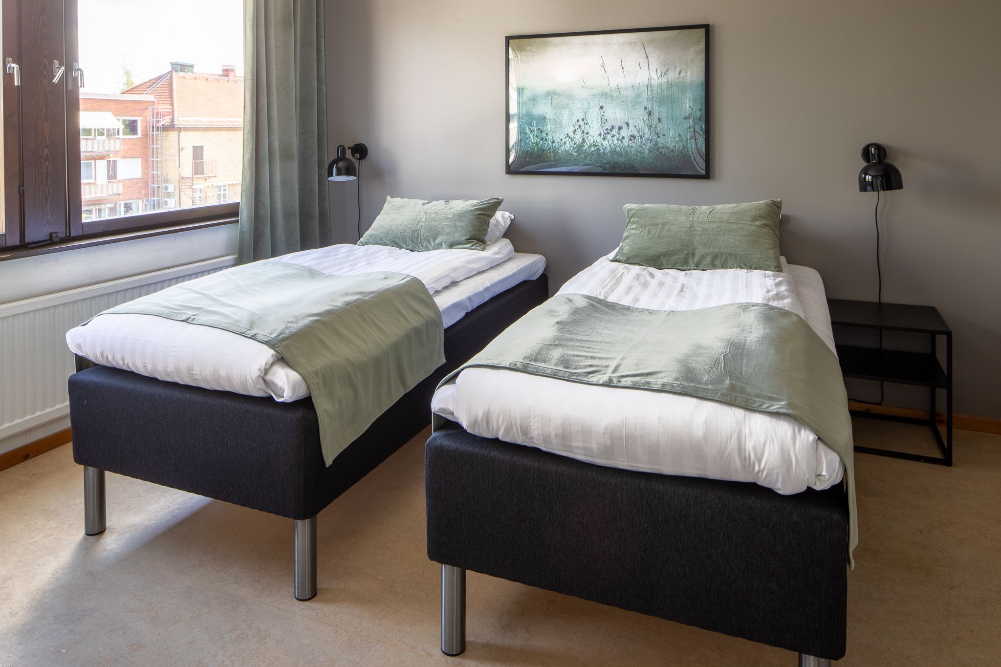 A room with two beds, a window with curtains, a painting on the wall, and a nightstand with a lamp.