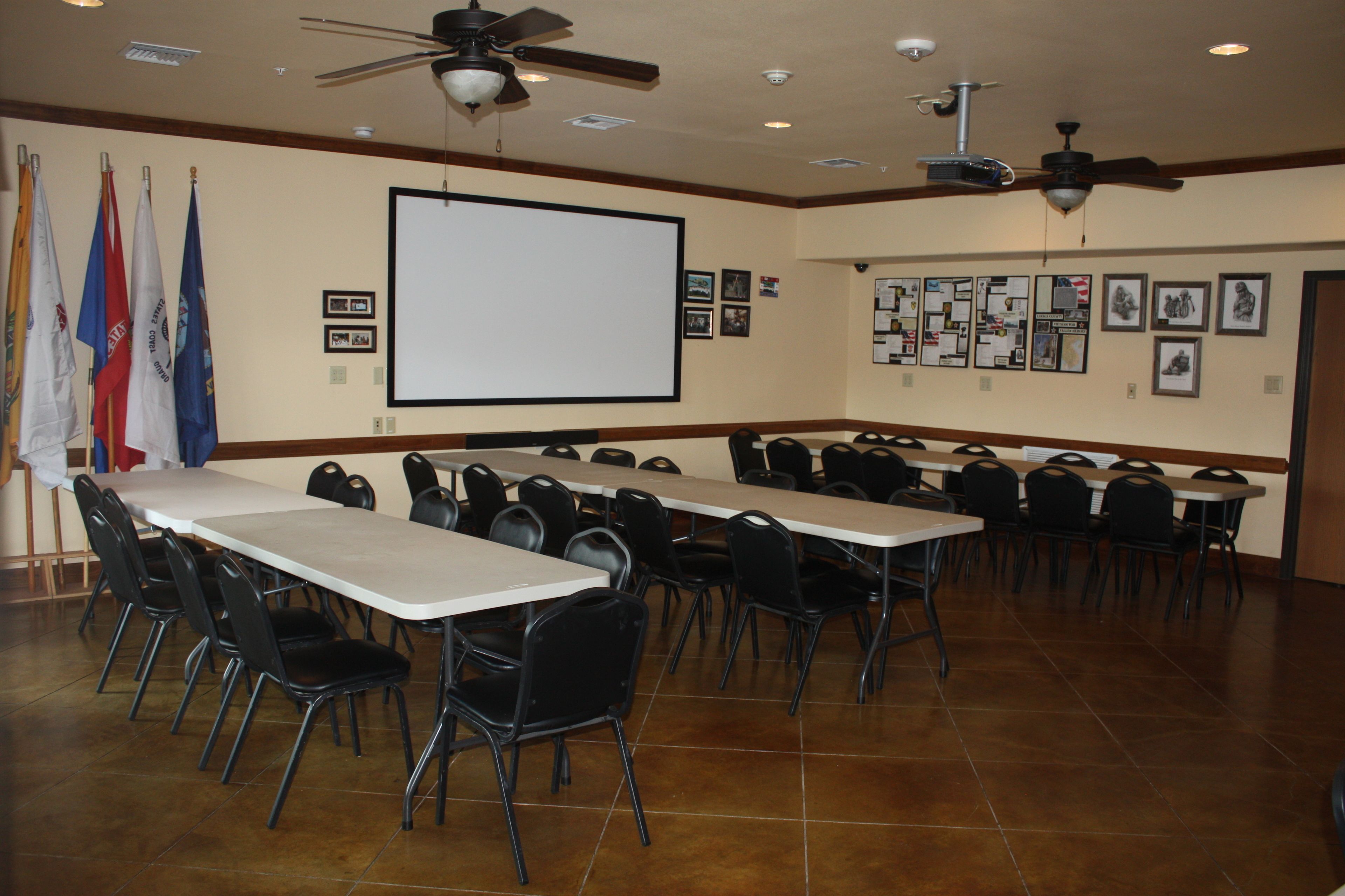A conference room with multiple tables and chairs, a projector screen, ceiling fans, and flags on the wall.
