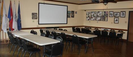 A conference room with multiple tables and chairs, a projector screen, ceiling fans, and flags on the wall.