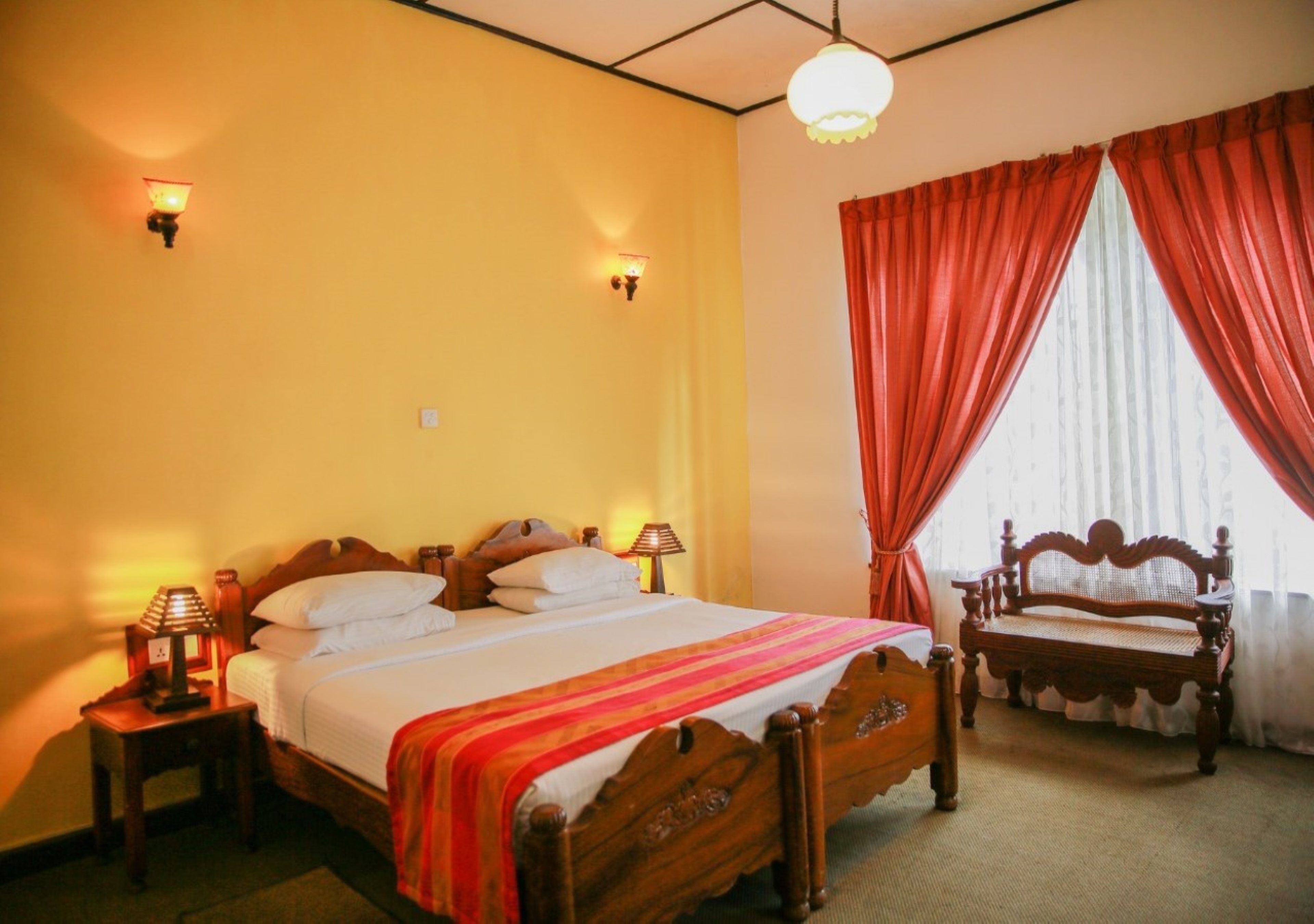A hotel room with a wooden bed, two bedside tables with lamps, a bench, and a large window with red curtains.