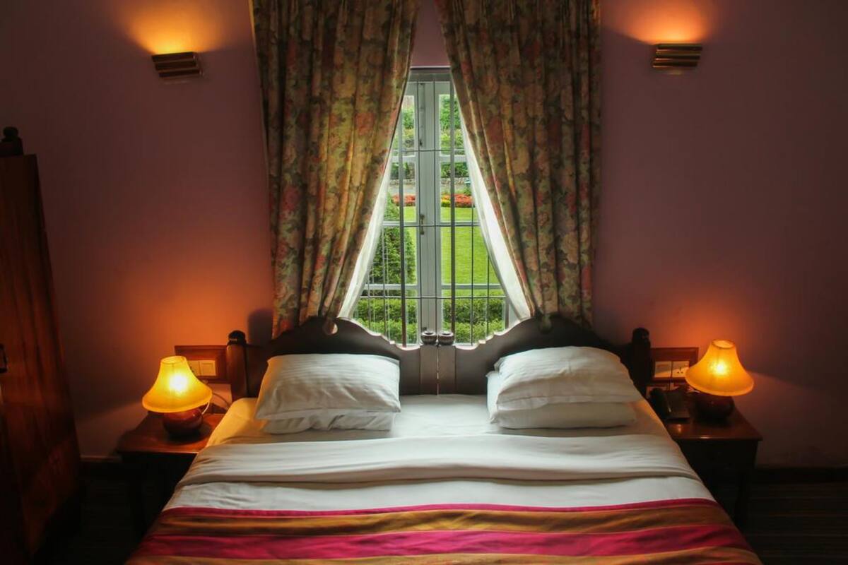 Een tweepersoonsbed met witte lakens, twee bedlampjes en een raam met gordijnen.