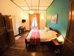 A hotel room with a bed, bedside table, lamp, desk, chair, and a TV on the wall. - Hotel Glendower (Nuwara Eliya)