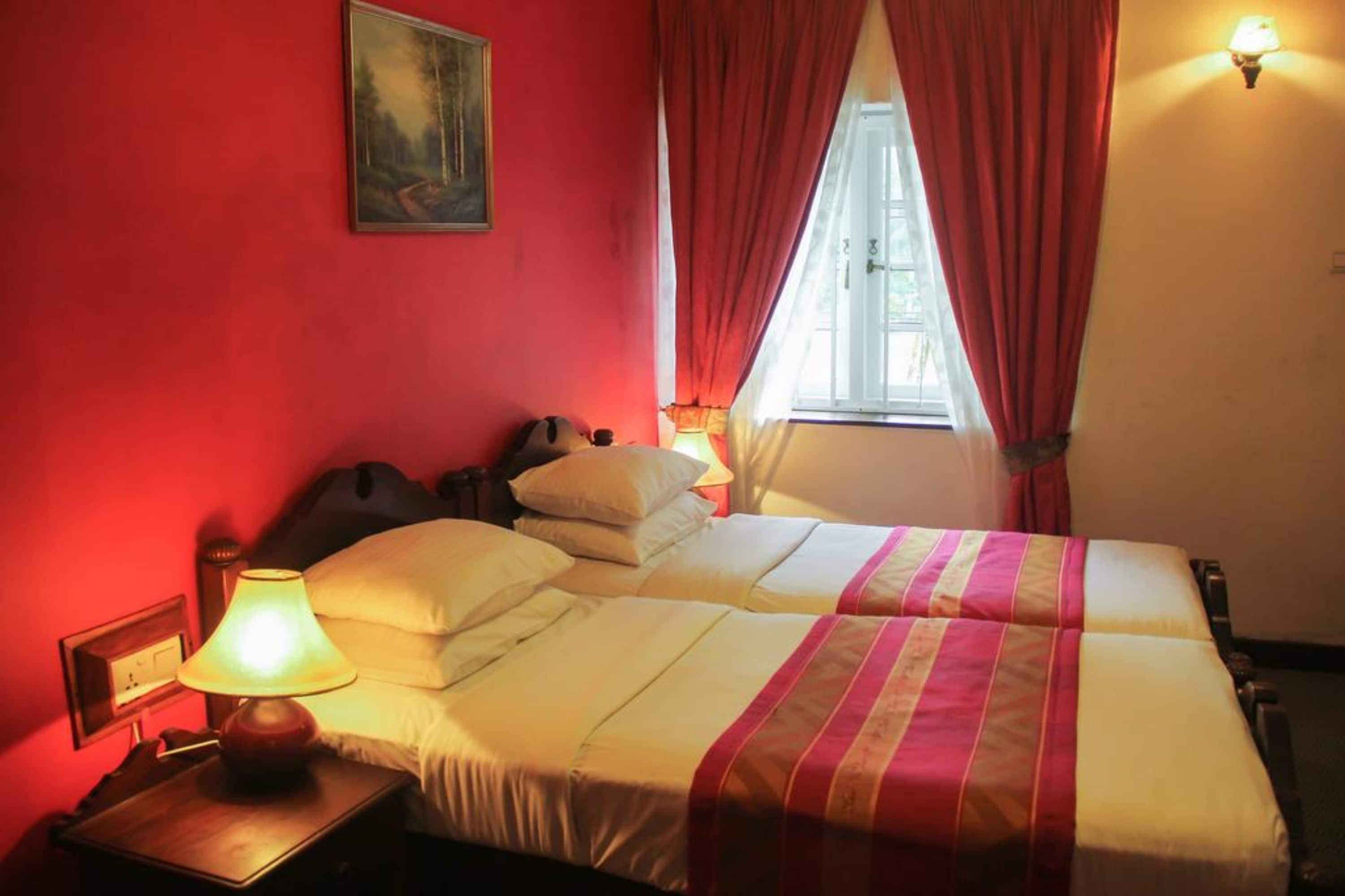 A hotel room with two beds, a nightstand with a lamp, a painting on the wall, and a window with red curtains.