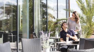 Een terras met rieten meubels, plantenpotten en een glazen wand met uitzicht op de omgeving.