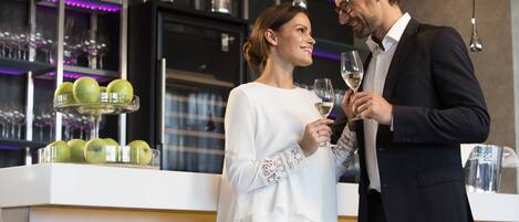 Una pareja está de pie en un área de bar moderna, cada uno con una copa de vino, una estantería con manzanas verdes y una botella de vino al fondo.
