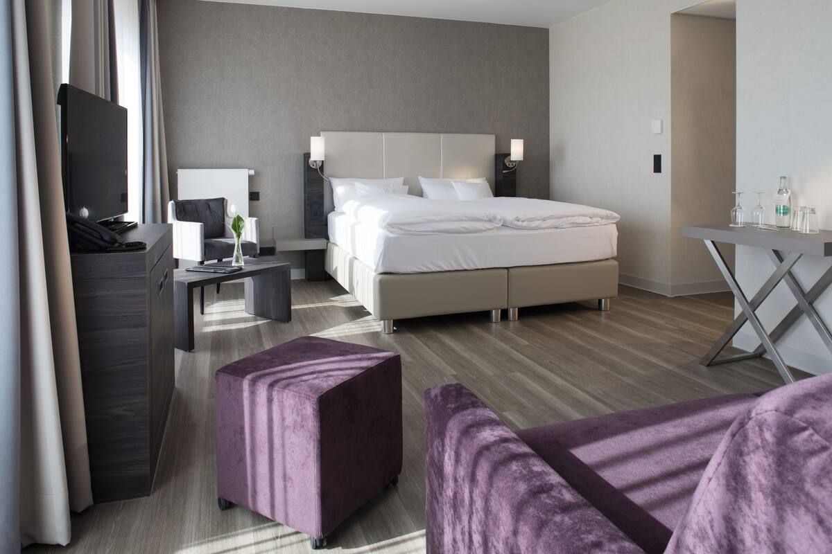 A modern hotel room with a large bed, a desk, a TV, and a seating area with purple ottomans.