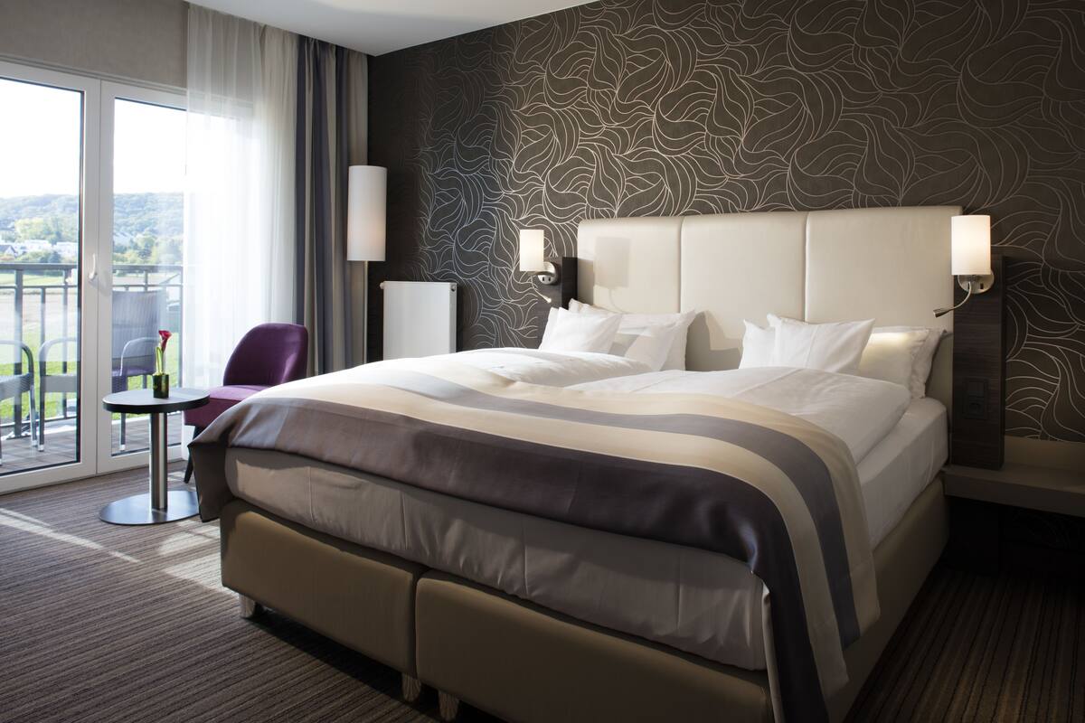 A hotel room with a large bed, two bedside tables, a chair, and a view of the outdoors.