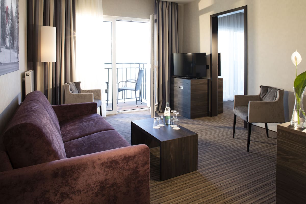 A modern hotel room with a sofa, armchairs, a coffee table, a TV, and a balcony with curtains.