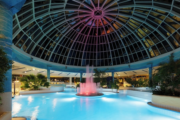 A large indoor swimming pool with a central fountain, surrounded by palm trees and a glass ceiling structure.
