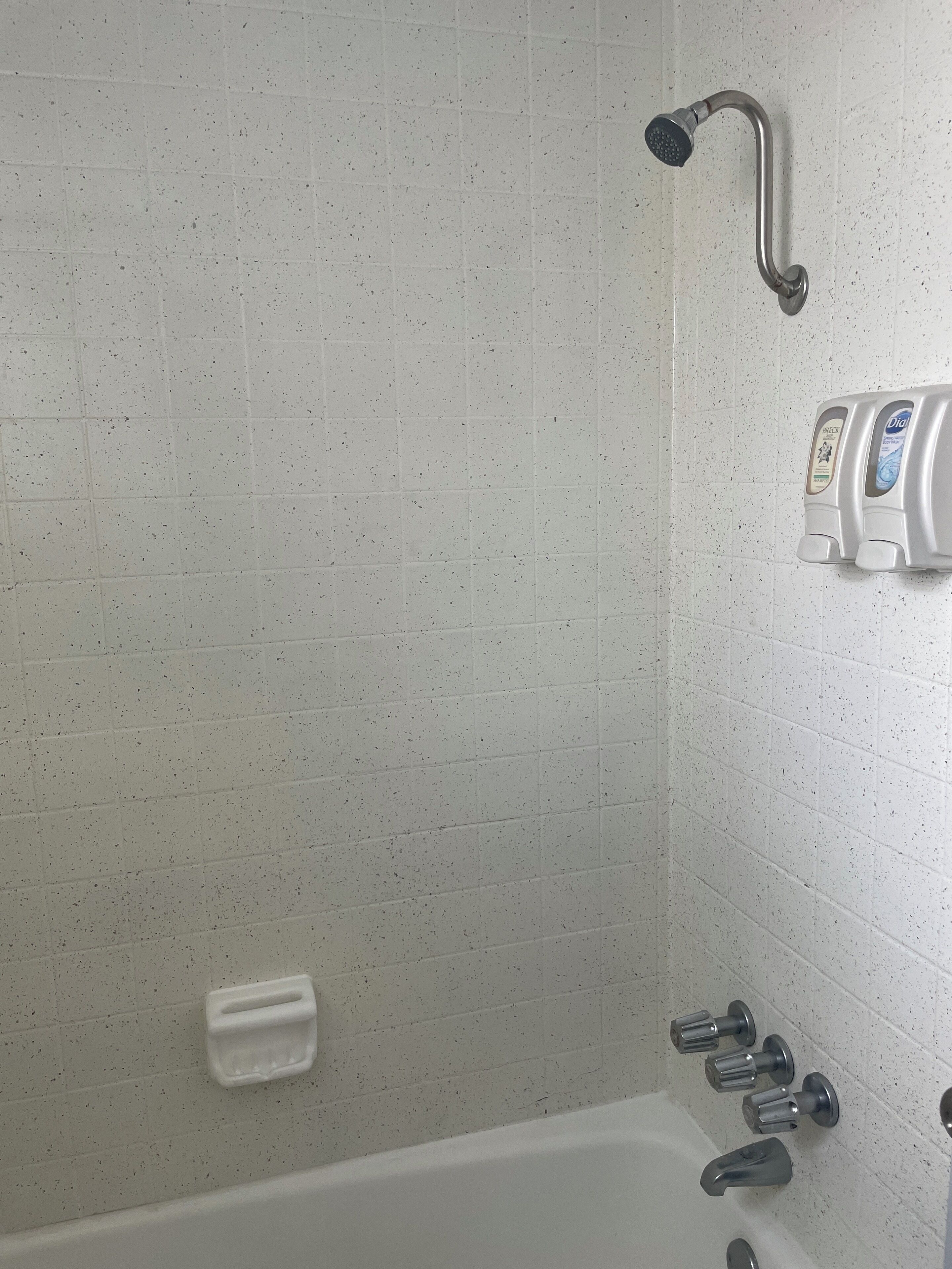 A bathroom with white speckled tiles, a showerhead, a soap dispenser, and a bathtub with multiple faucets.
