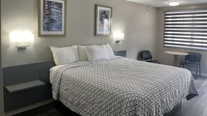 A hotel room with a bed, two wall-mounted lights, a window with blinds, and two framed pictures on the wall. - Economy Inn (St. Augustine)