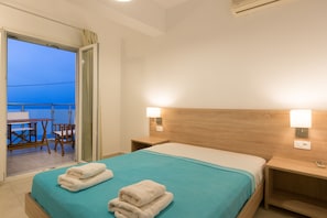 A hotel room with a bed, two bedside tables, a nightstand, a window with curtains, and a view of the outside balcony. - Socrates Seaside Apartments (Agios Nikolaos)