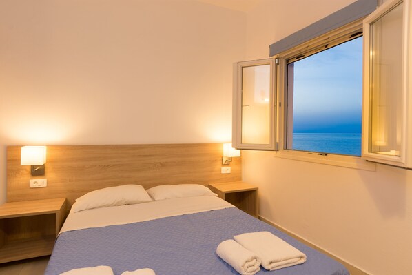 A hotel room with a bed, two bedside lamps, a window with a view of the sea, and a nightstand. - Socrates Seaside Apartments (Agios Nikolaos)