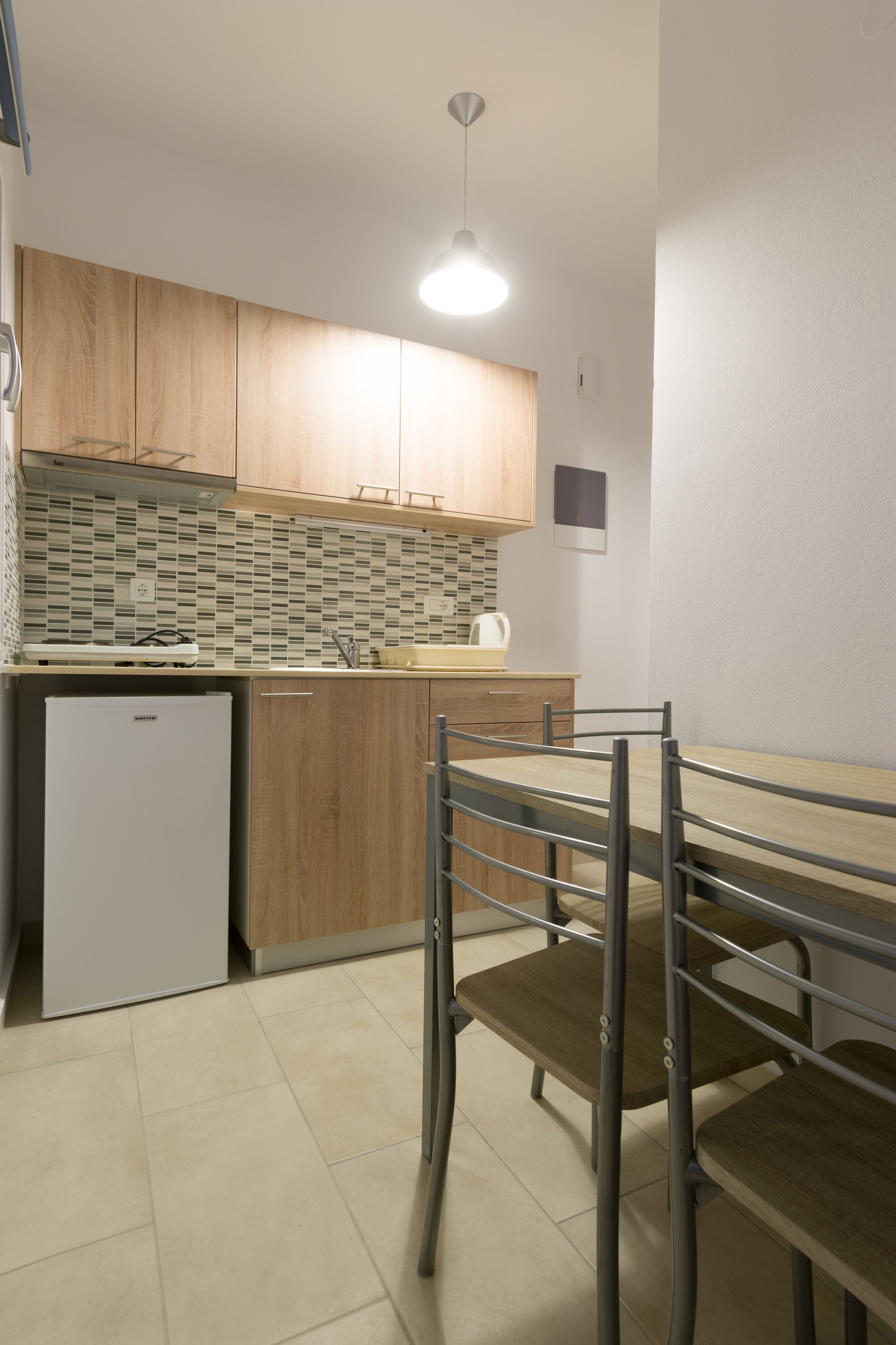Une cuisine compacte avec des meubles en bois, un petit réfrigérateur, un plan de travail carrelé et une table à manger en métal avec des chaises.