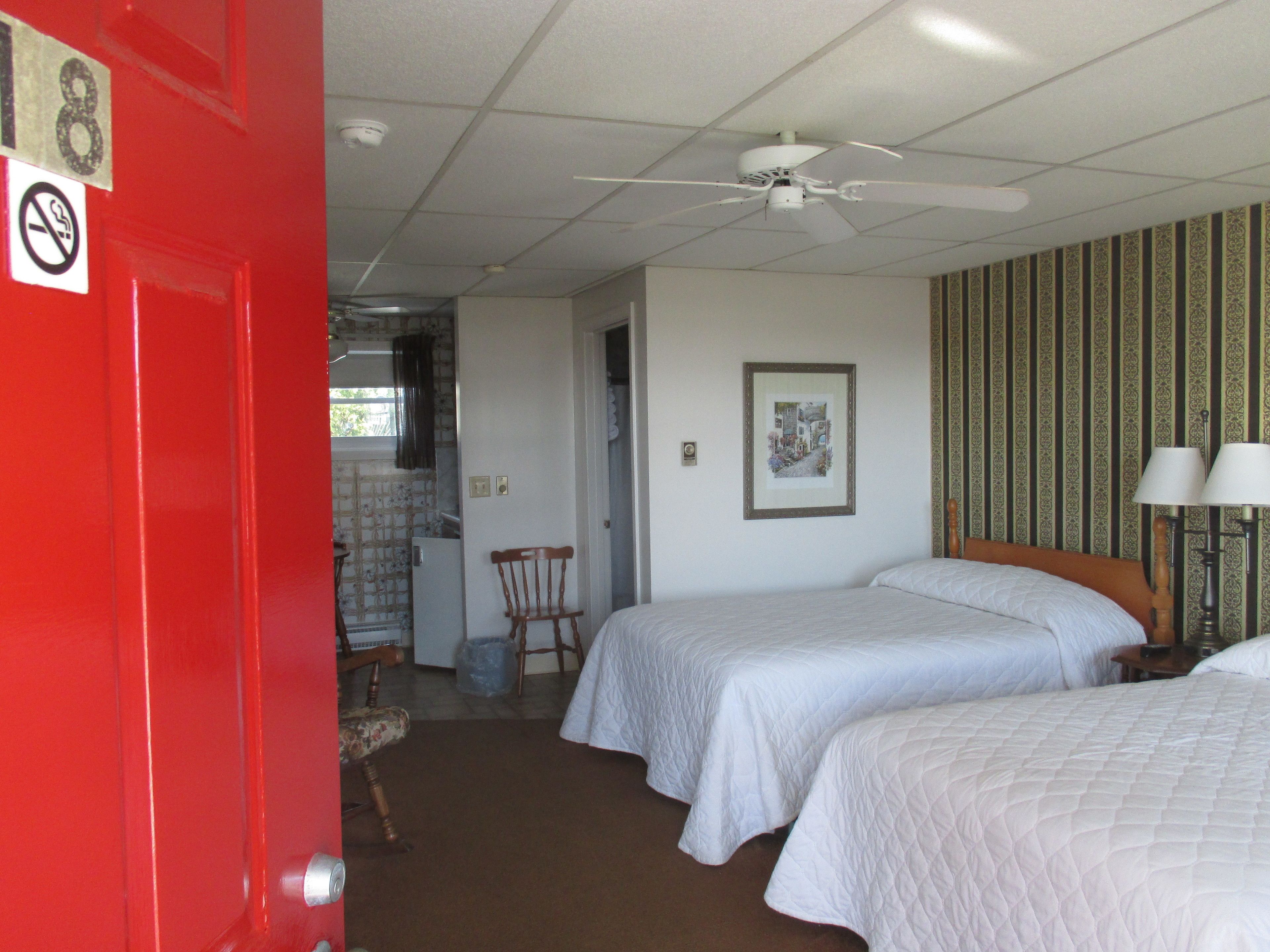 Une chambre d’hôtel avec deux lits, un ventilateur de plafond, un tableau encadré au mur et une porte rouge avec un panneau « interdiction de fumer ».