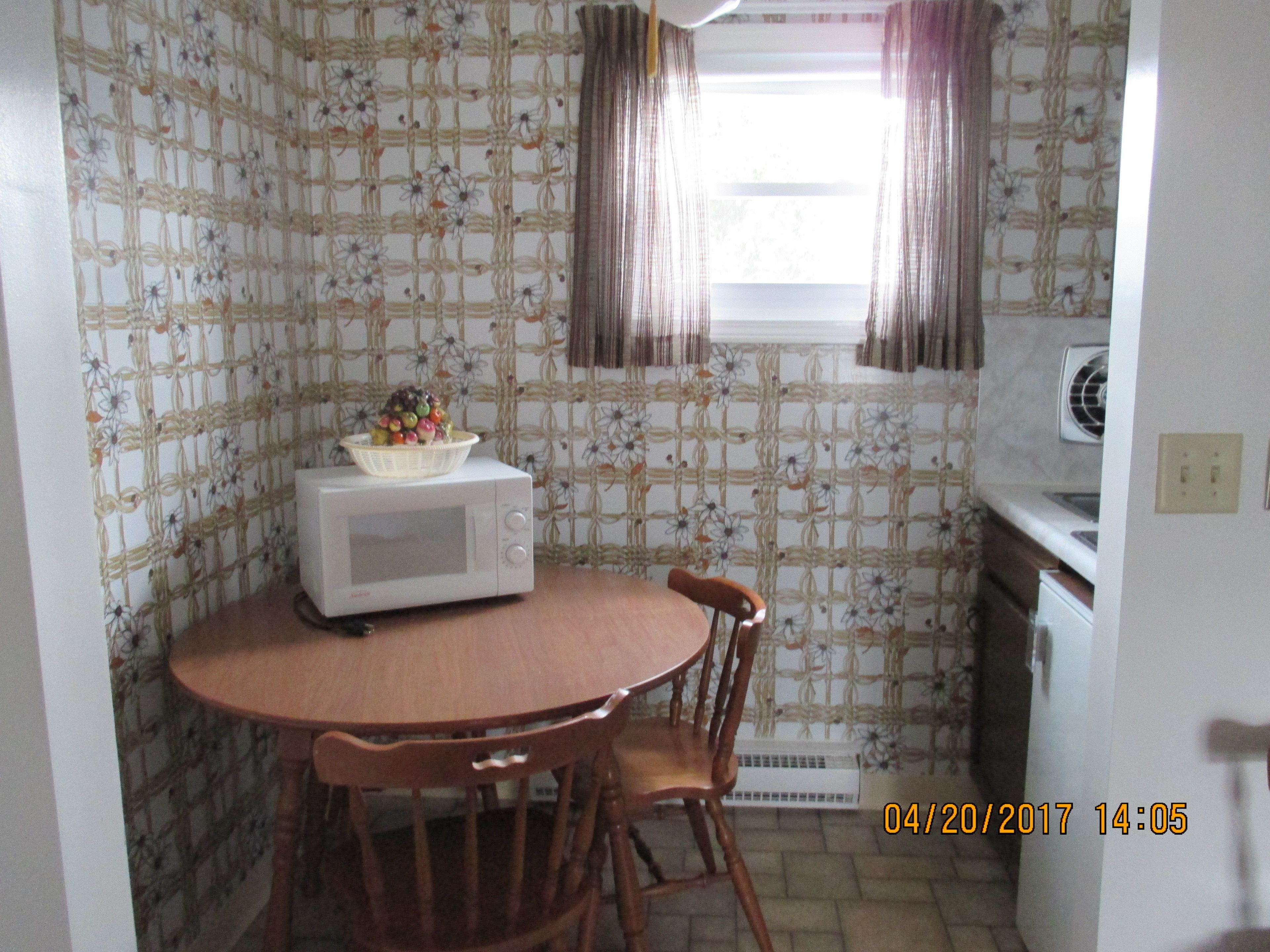 A small kitchen with a microwave, a wooden table, and chairs. There is a window with curtains, and a wall with a floral pattern.
