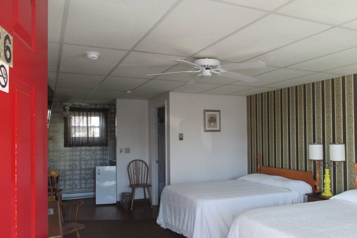 Ein Hotelzimmer mit zwei Betten, einem Deckenventilator, einem kleinen Schreibtisch mit Stuhl, einem Kühlschrank und einem Fenster mit Vorhängen.