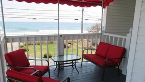 A covered outdoor seating area with red chairs and a small table, overlooking a grassy area and the ocean. - Long Beach Motor Inn (York Beach)