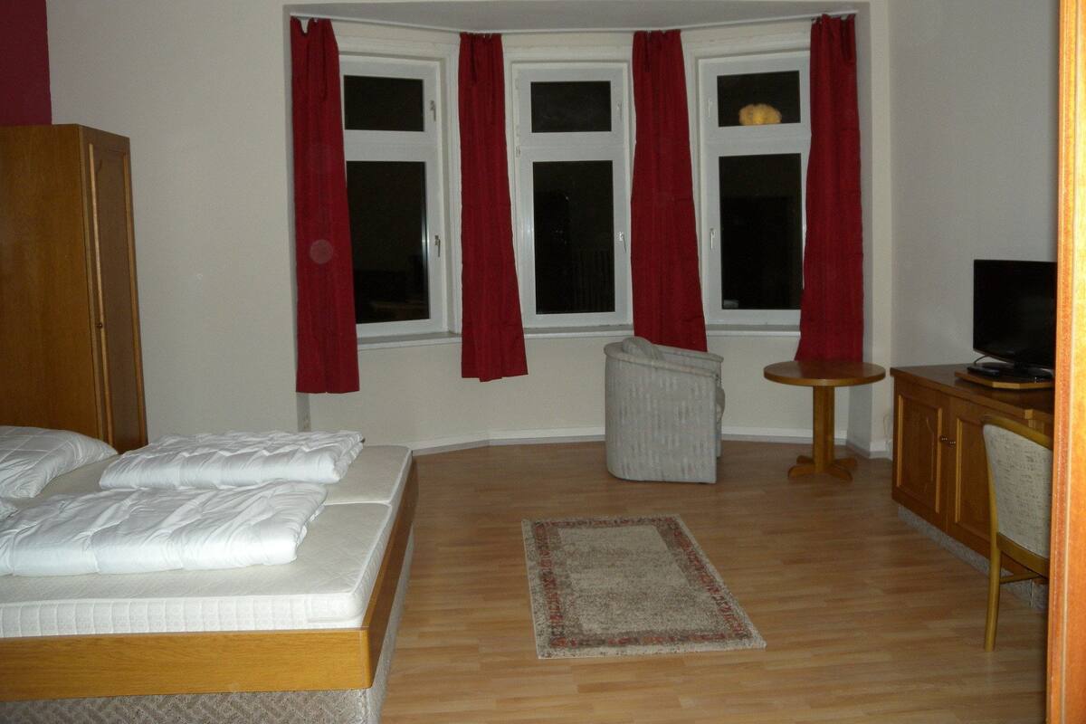 A bedroom with a bed, a small armchair, a television, and a window with curtains.