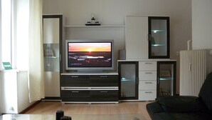 A modern living room with a flat-screen TV on a stand, a glass coffee table, a black leather sofa, and a cabinet with glass doors.