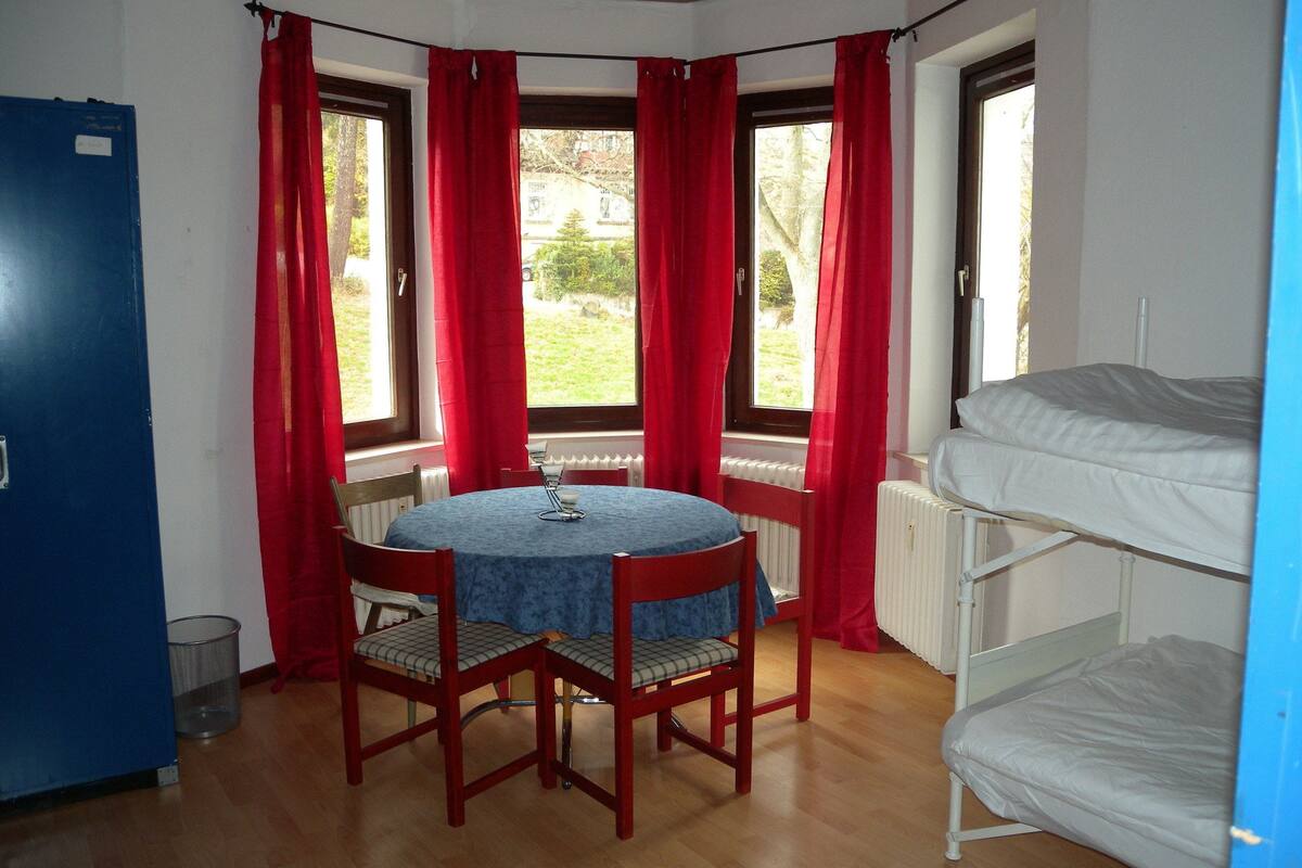 A room with a round table, two chairs, a bunk bed, and a window with red curtains.