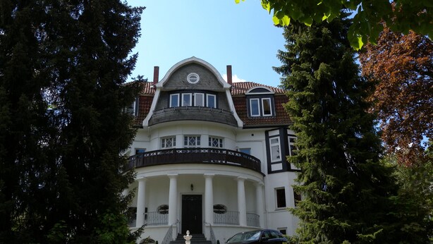 A large, two-story building with a symmetrical facade, a central entrance with steps, and a fountain in front.
