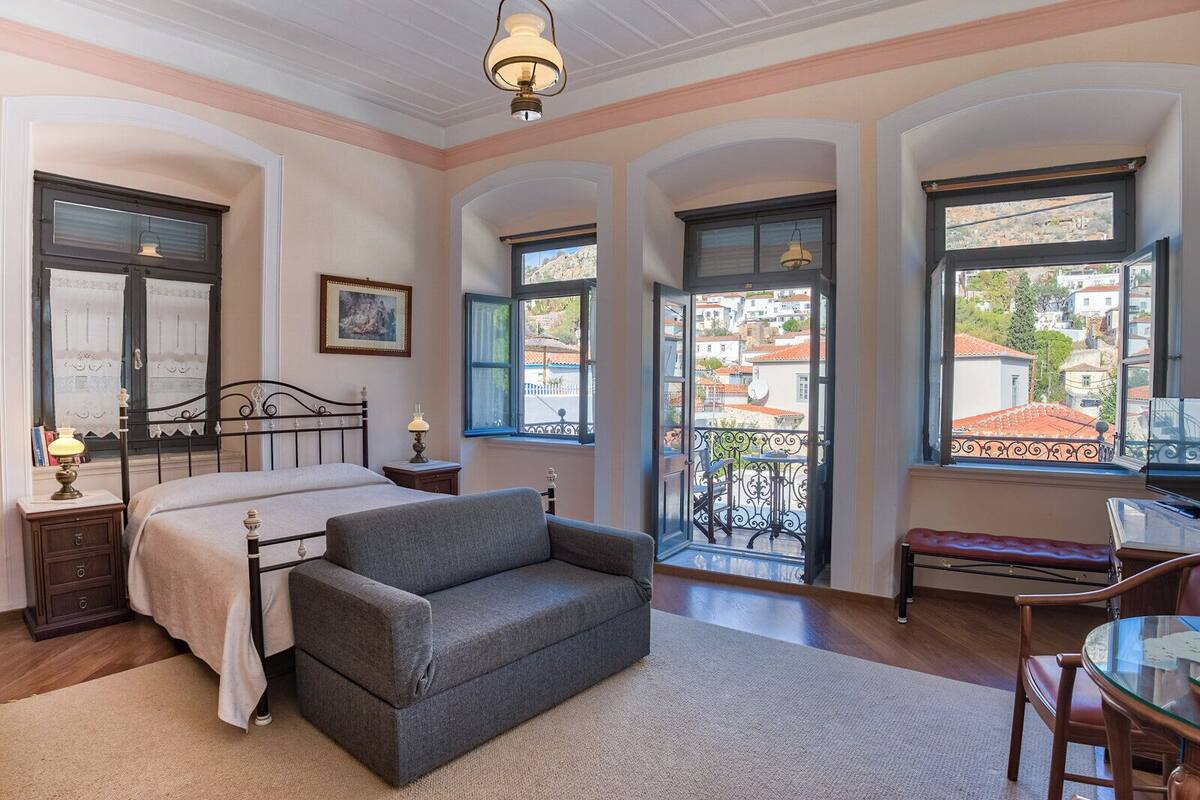 A bedroom with a bed, a grey sofa, a small table with a lamp, a chair, and a balcony with a view of the city.