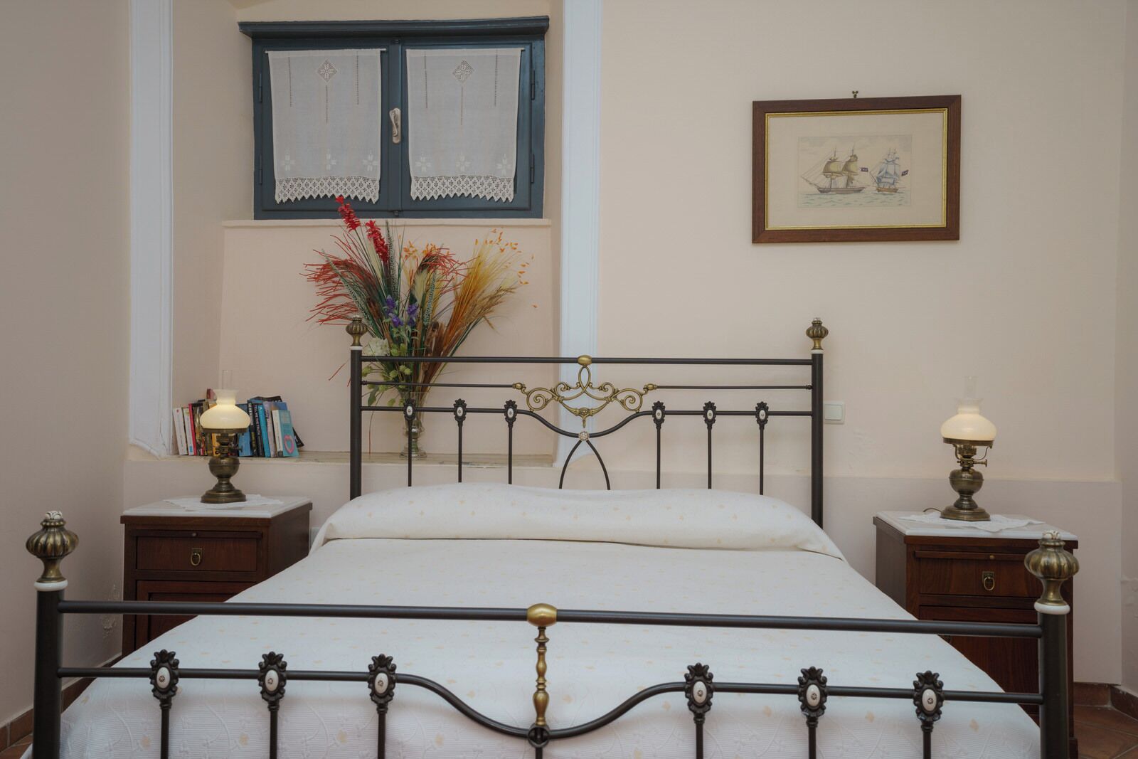 A bedroom with a metal-framed bed, two bedside tables with lamps, a window with lace curtains, a framed picture on the wall, and a vase with flowers on the windowsill.