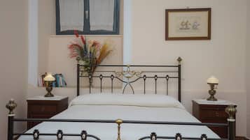 A bedroom with a metal-framed bed, two bedside tables with lamps, a window with lace curtains, a framed picture on the wall, and a vase with flowers on the windowsill.