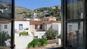 A view from a window of a coastal town with buildings on a hillside, including a white building with a blue door and a balcony with plants.