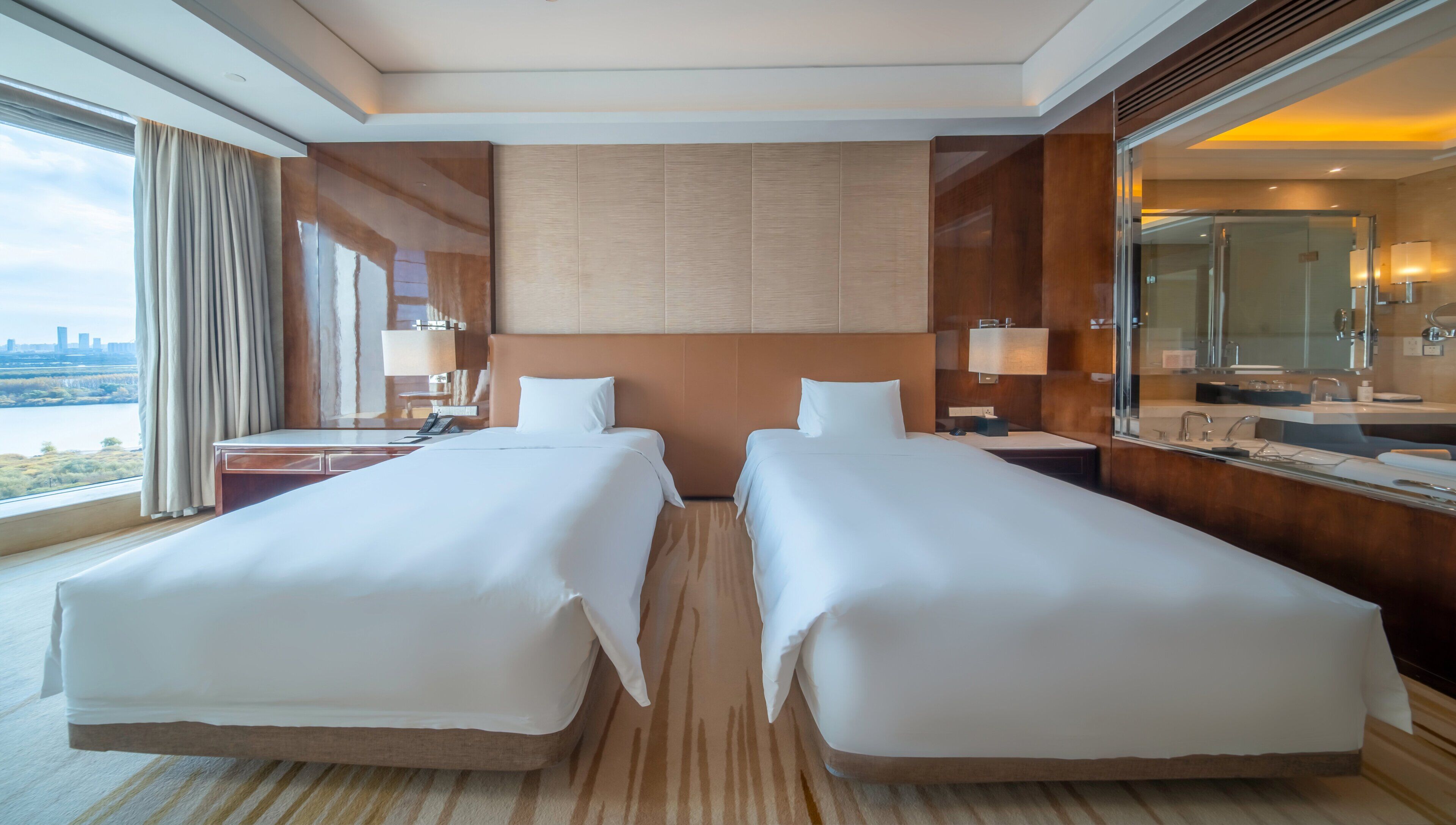 Habitación de hotel con dos camas, un baño con espejo y una mesita de noche con lámpara, y un ventanal con vistas.