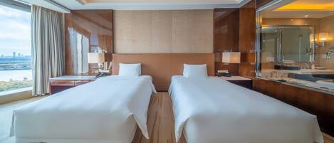 Habitación de hotel con dos camas, un baño con espejo y una mesita de noche con lámpara, y un ventanal con vistas.