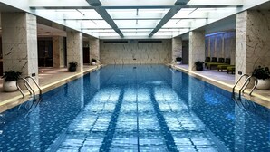 Una amplia piscina interior con agua cristalina y azul, rodeada de paredes y columnas de mármol, y con varios puntos de acceso.