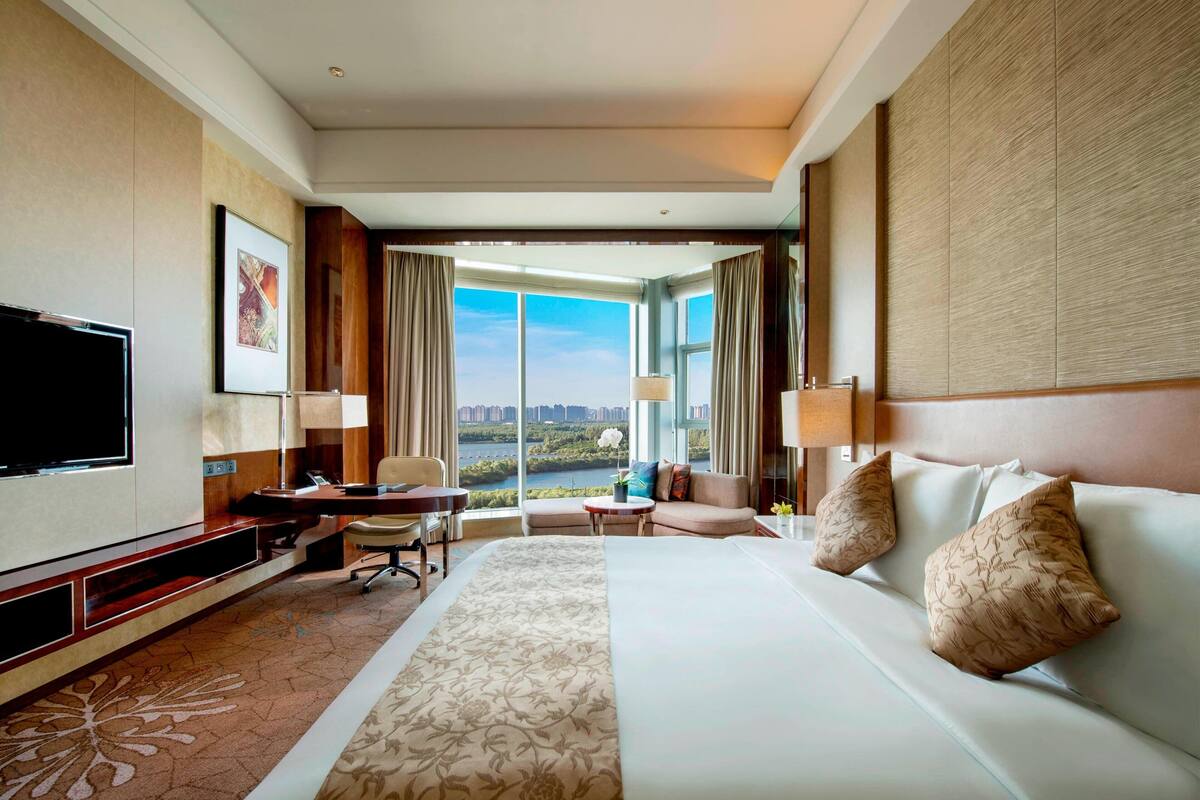 A hotel room with a large bed, a desk, a chair, a TV, and a view of the city.