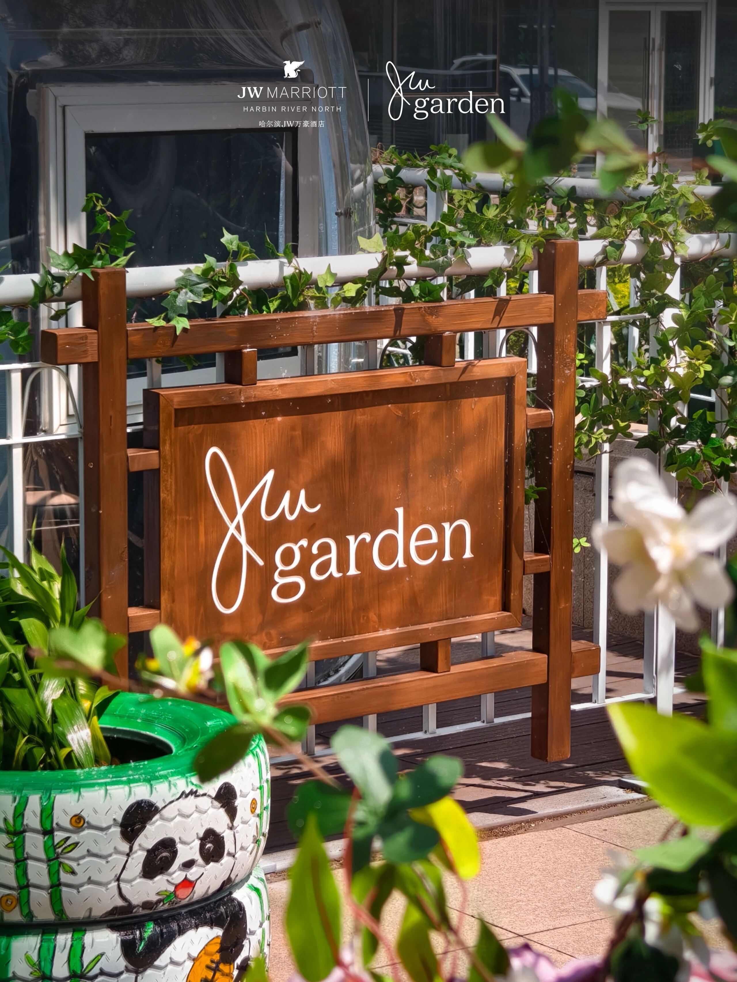 Een houten bordje met de tekst 'Juw Garden', omringd door groen en een plantenpot met panda-thema.