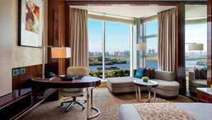Habitación de hotel con escritorio, sofá, una mesita y cama. Se aprecia una ventana grande con vistas a un paisaje urbano y una masa de agua.