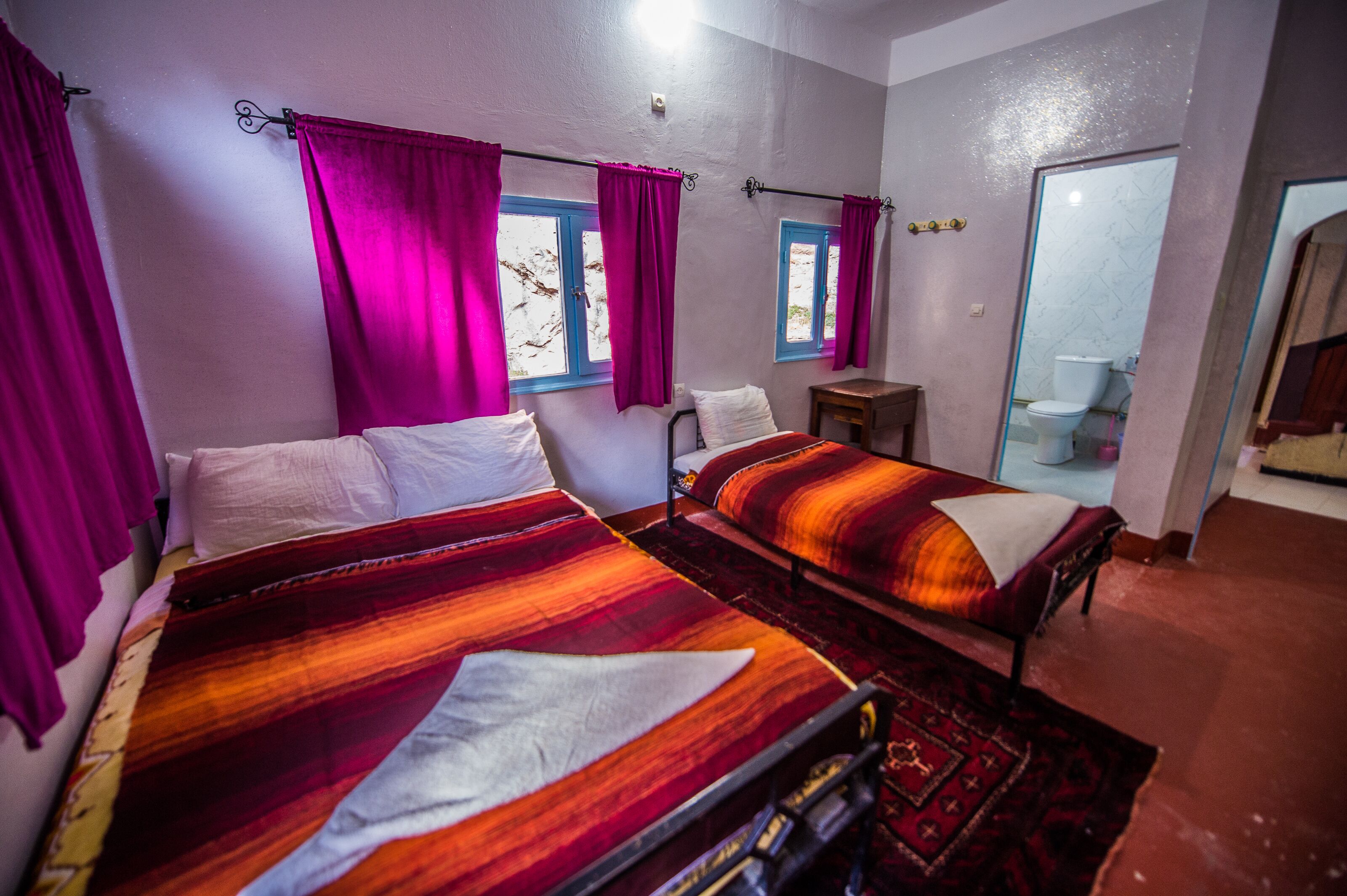 Habitación con dos camas, cortinas moradas, una mesita pequeña y baño visible.