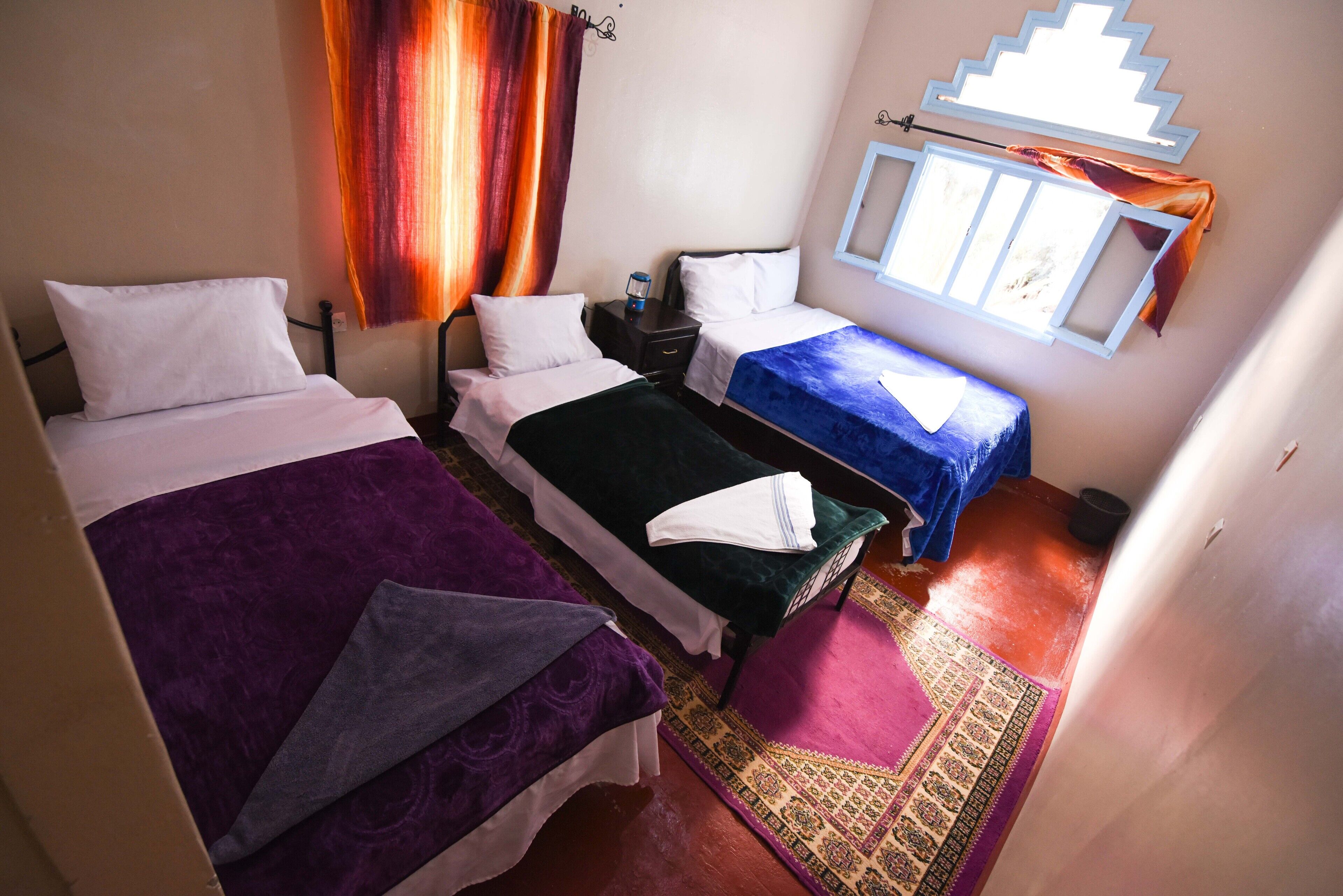 A room with two beds, a window with curtains, a small table with a lamp, and a patterned rug.