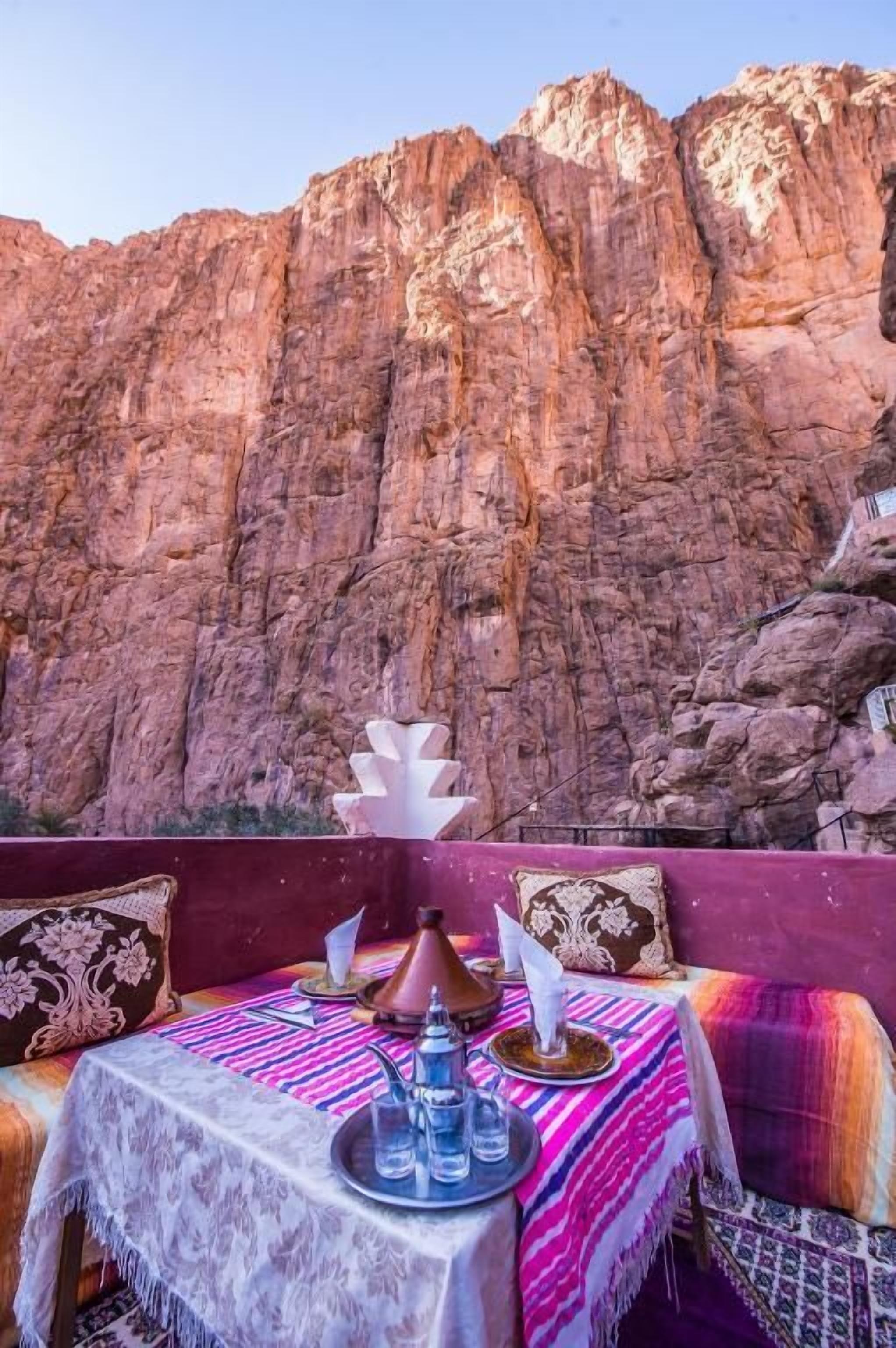 Un coin repas avec une table dressée pour deux, un tajine marocain, et une vue sur une montagne rocheuse.