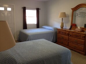 A bedroom with two beds, a dresser, a lamp, and a window with curtains. - McGregor Inn Motel (Gansevoort)
