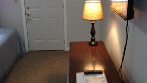 A hotel room with a wooden nightstand, a lamp, a framed picture on the wall, and a door. - McGregor Inn Motel (Gansevoort)
