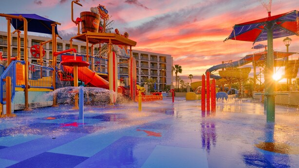 Een waterpark met een speelstructuur, glijbanen en een zonsondergang op de achtergrond.
