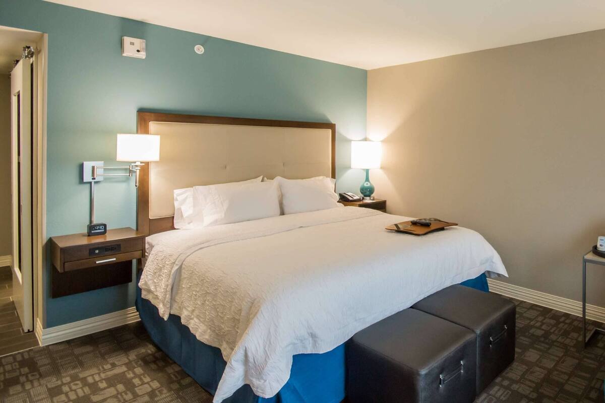 A hotel room with a bed, bedside table, lamp, and a blue armchair with a patterned pillow.