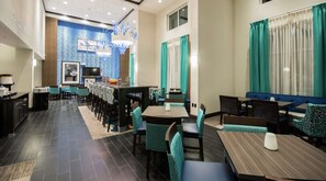 A restaurant with a long counter, multiple tables, and chairs. There is a television mounted on the wall and a window with curtains. - Hampton Inn & Suites Orlando At SeaWorld (Orlando)