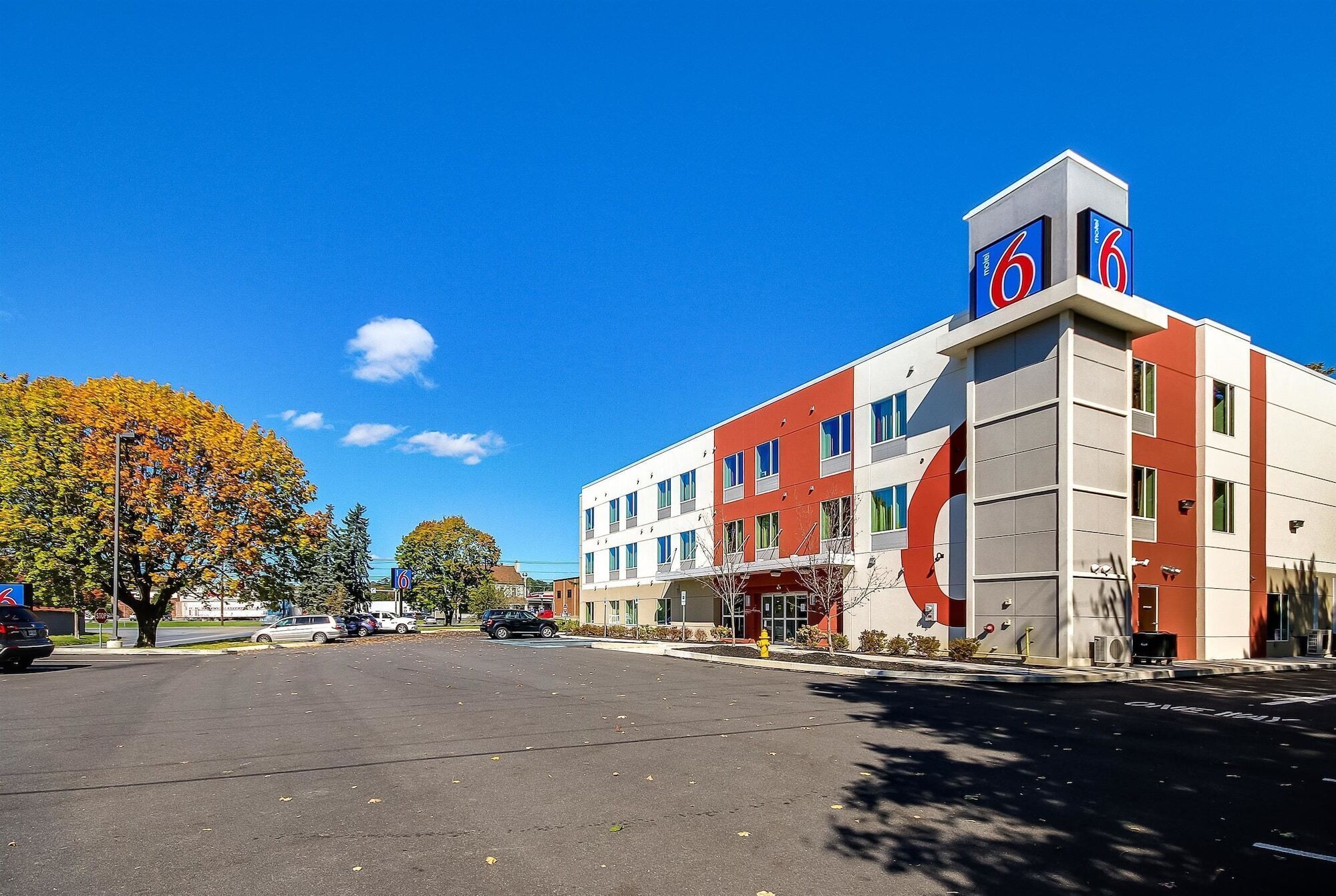 Un motel de diseño moderno, con un llamativo letrero con el número '6' en el techo, un estacionamiento y árboles de colores otoñales al fondo.